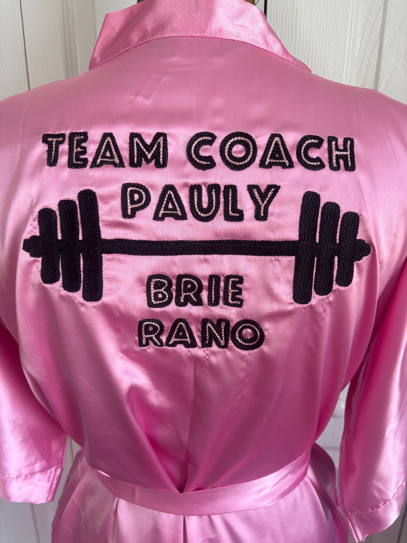 Custom commissioned chainstitch embroidered pink satin robe for a bodybuilding competition. Shows "Team Coach Pauly" in pink and black ryaon thread, a barbell, and the bodybuilder's name at the bottom.