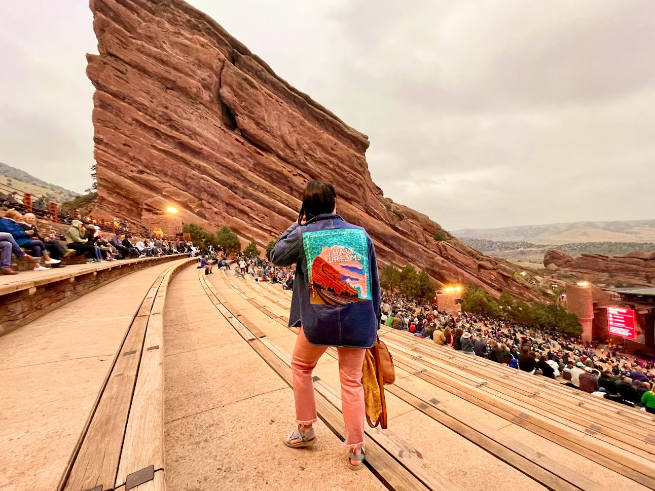 woman at red rocks stadium wearing a custom chainstitched denim jacket stitched with a sunset, red rocks, and text that says "I Have Dreams" lyrics inspired by Brandi Carlile.